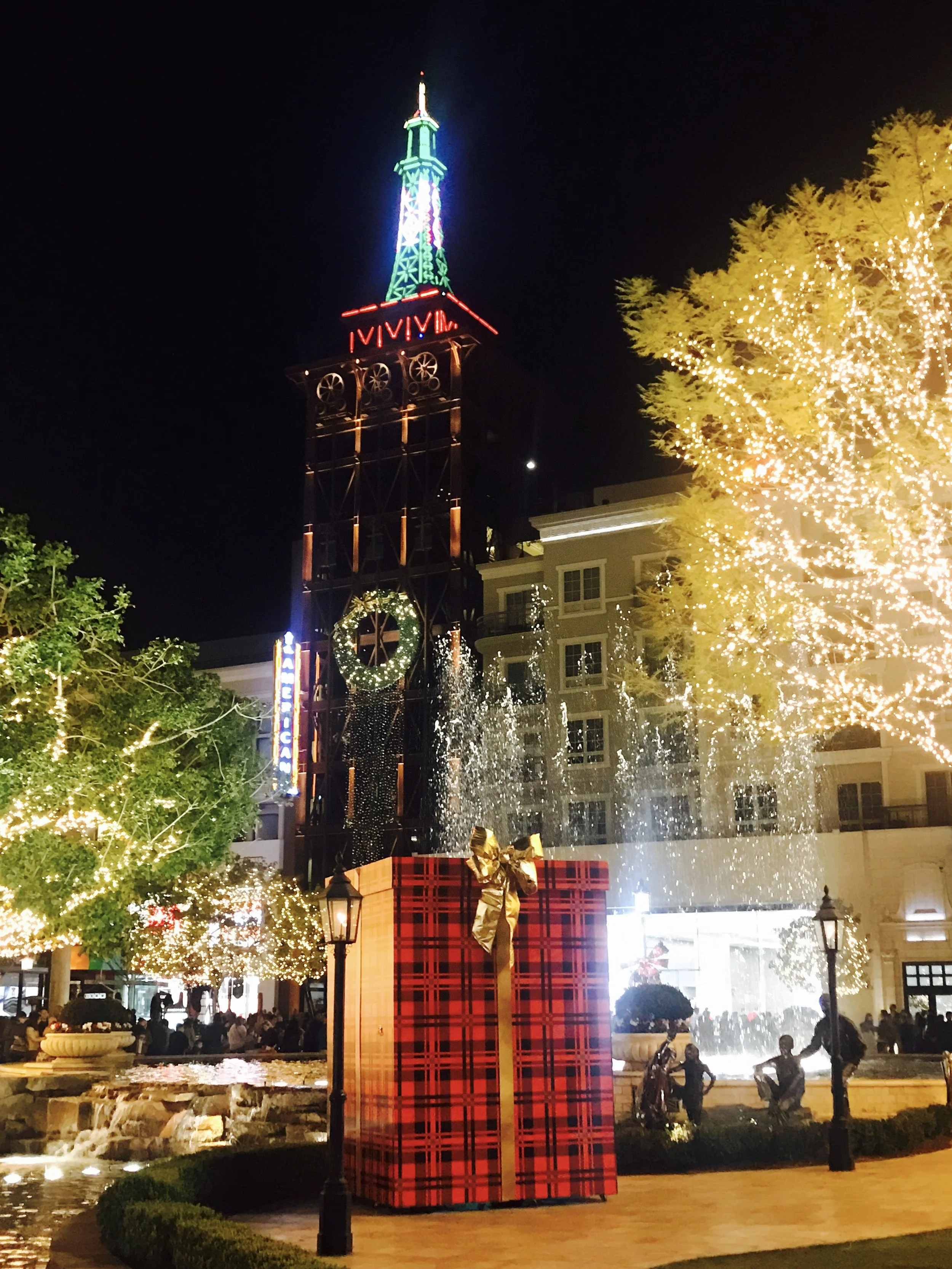 Nighttime holiday scene at The Americana at Brand with lit tower, fountain and giant plaid gift box
